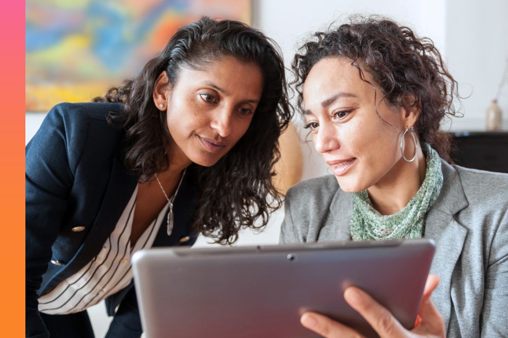 A woman sitting down while holding a tablet and showing another woman