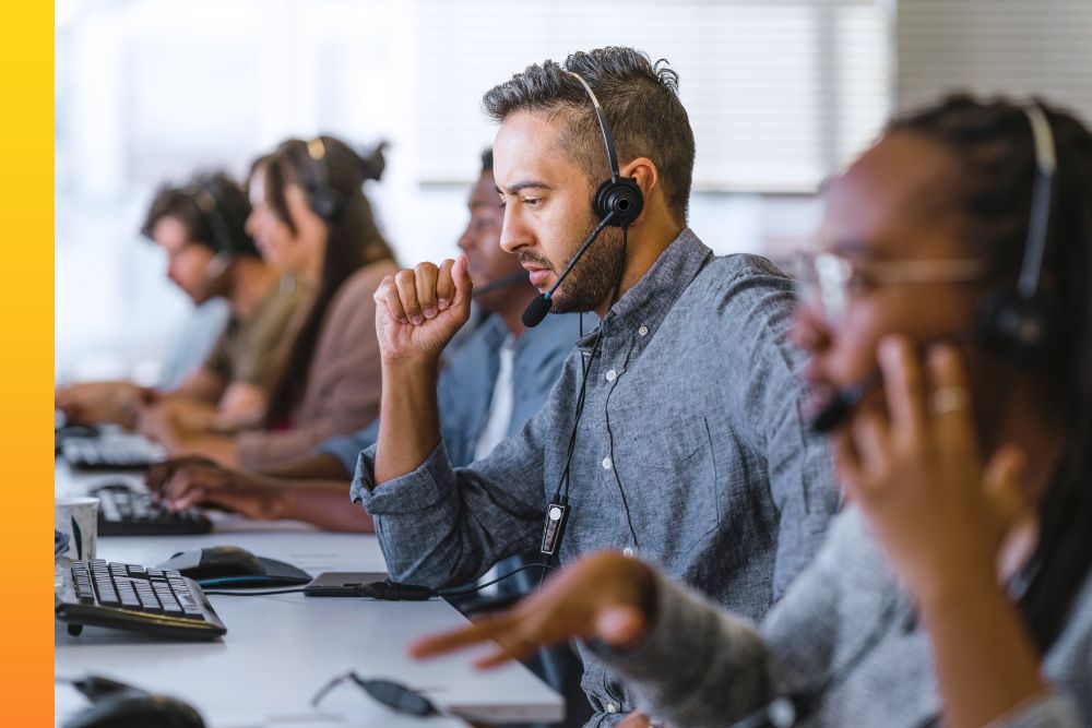 Man in a grey shirt and headset sitting in a row of computers with several other people 