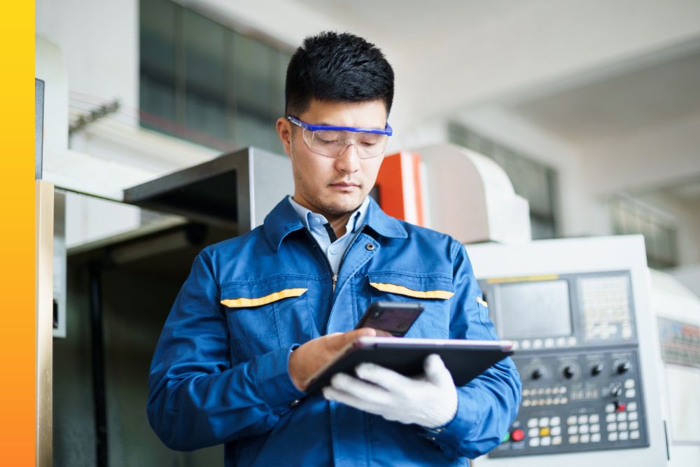 Man in blue coveralls and safety glasses while standing and working on a tablet device