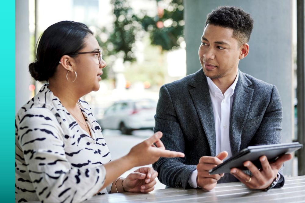 Man in a grey jacket sitting across from a woman in a white shirt while looking at a tablet