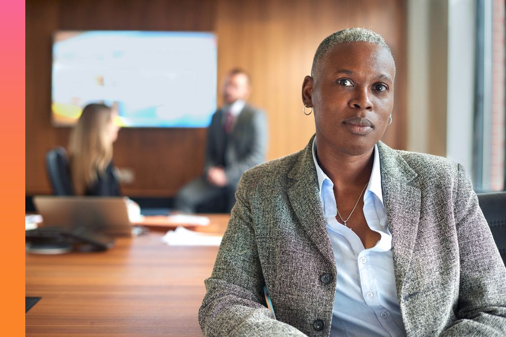Woman in a tan jacket sitting at a conference table while other people work in the back
