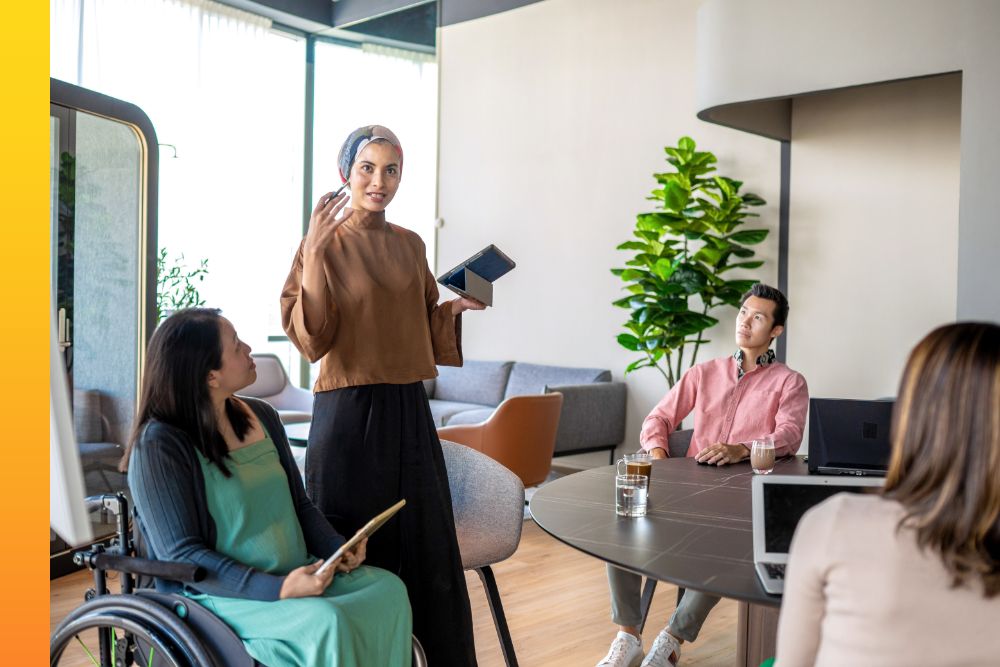 One person standing while holding a tablet and talking to three other people sitting down 