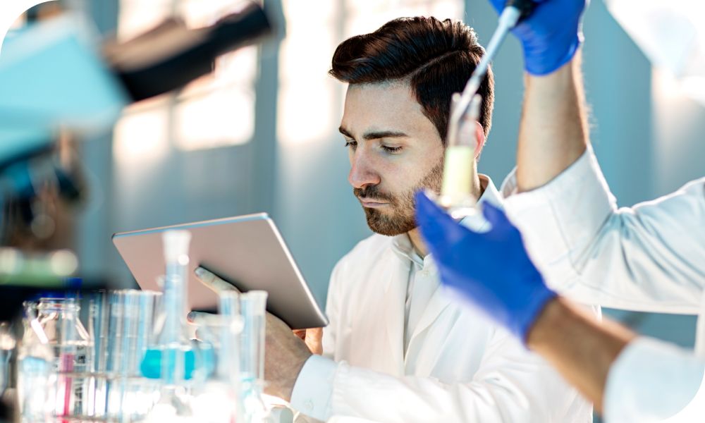 A pair of arms filling a beaker while a man in a white doctors coat sits behind and looks at a tablet