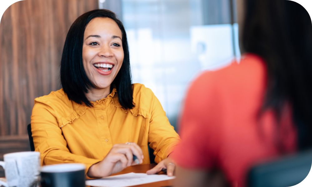 Woman in a light orange shirt sitting at a desk while talking to another person across from her