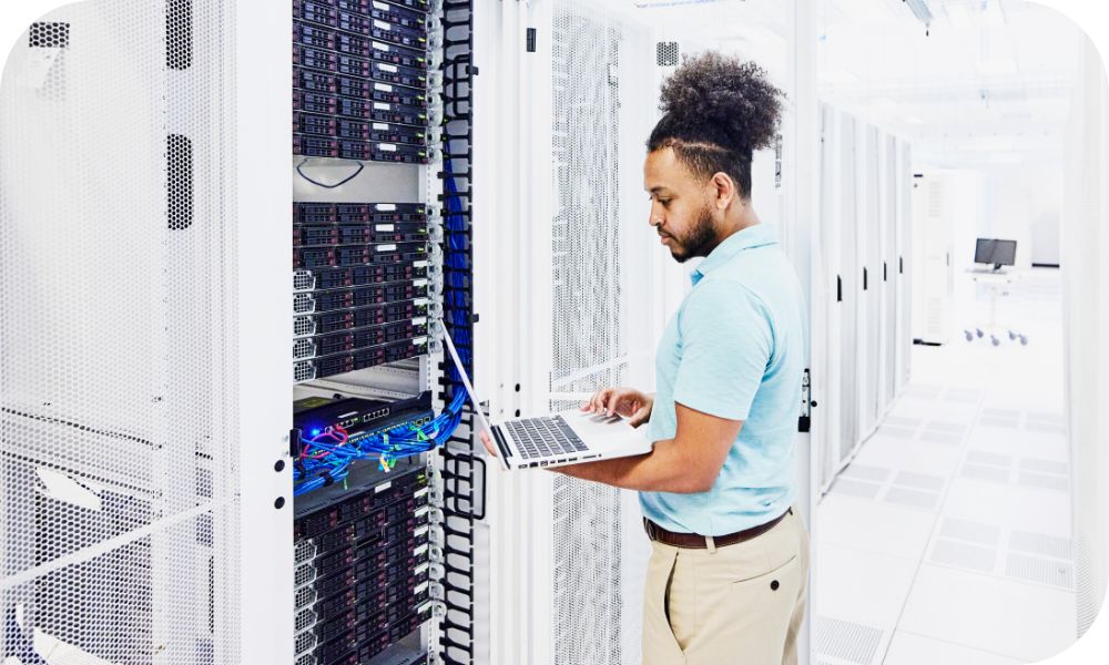 Man in a light shirt standing while holding a laptop in front of a server stack