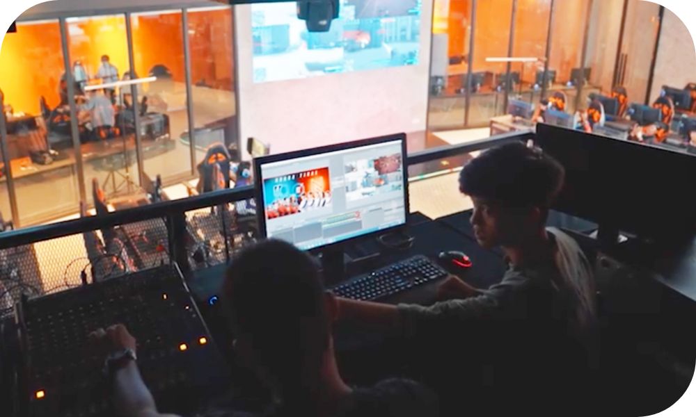 Two people in an elevated production booth working on a soundboard and computer above a gym floor