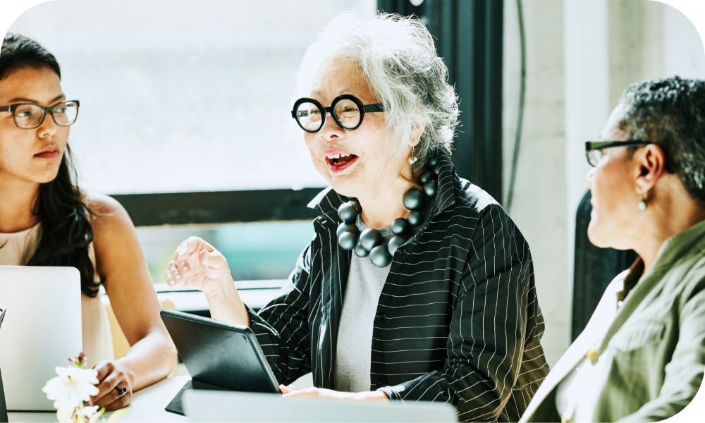 Woman in a black striped jacket sitting with two other people in front of laptops while talking