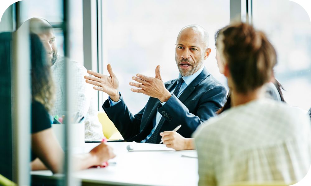 Man in a dark grey suit sitting across from other people at a conference table while talking