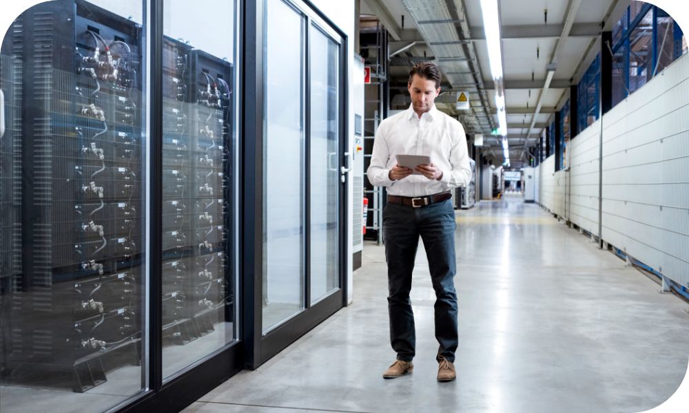 Man standing in a white shirt while holding a tablet next to a server stack