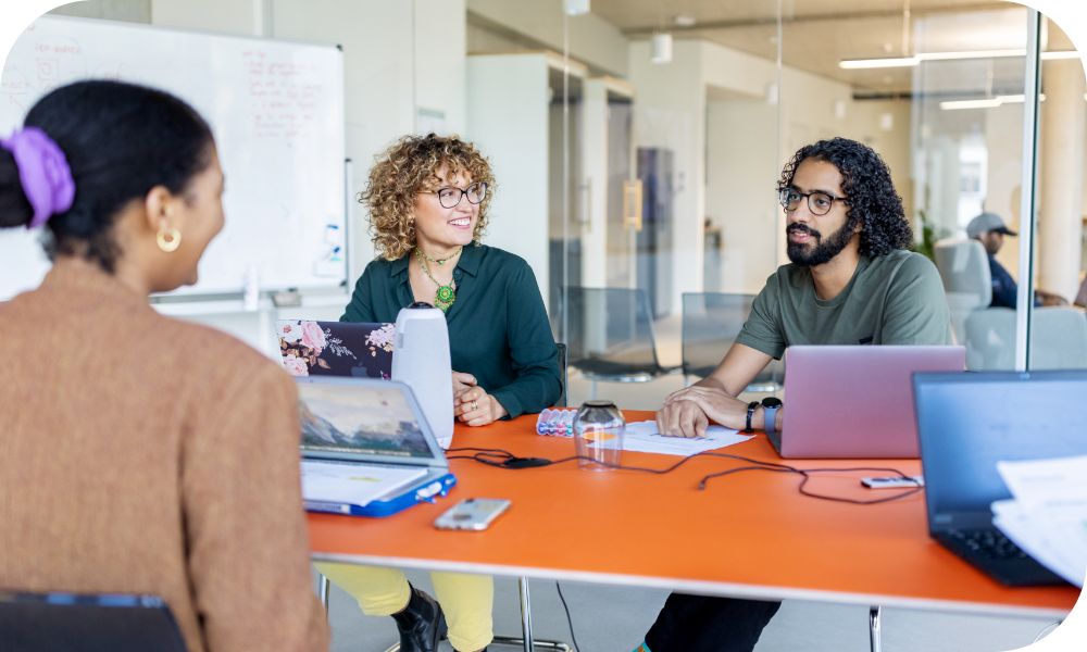 three people sitting across from each other at an orange conference table with laptops and papers on it
