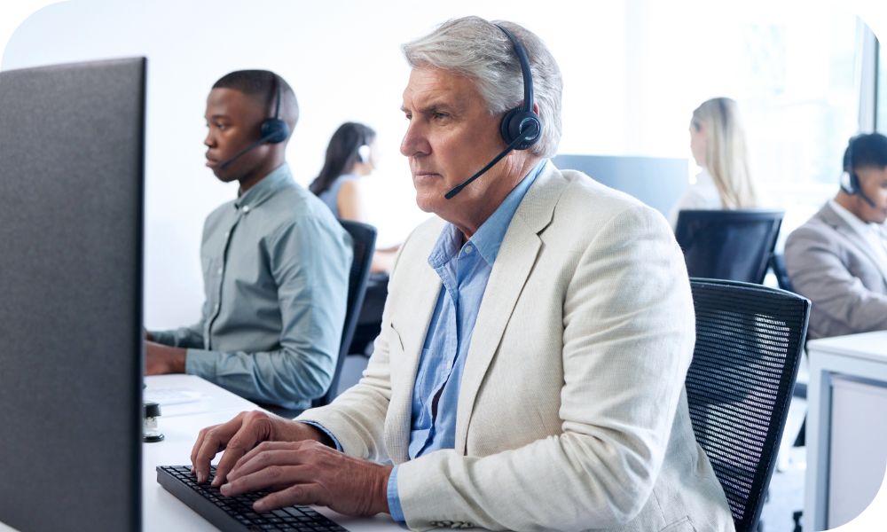 Man in white jacket and headset sitting while working on a computer while other people work in the background 