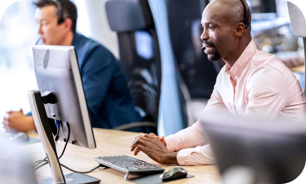 Man sitting in front of a computer while talking on a headset while another person is working in the background