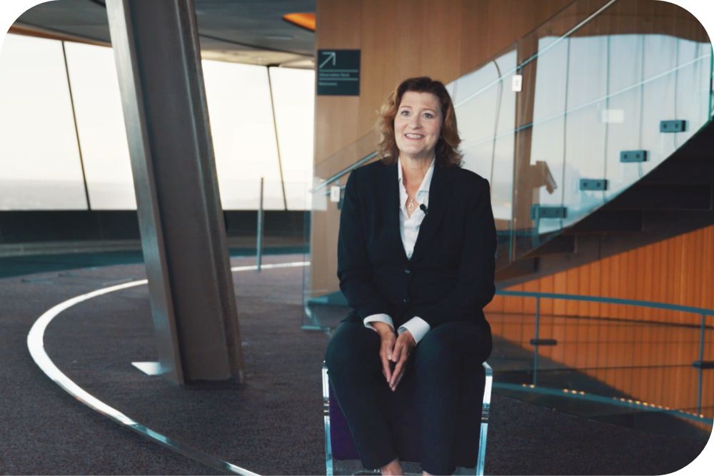 Karen Olson, Chief Operating Officer of the Seattle Space Needle, smiles while seated indoors near large windows and a modern staircase with glass railings.
