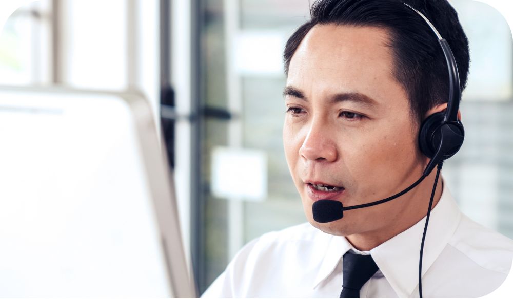A businessperson in a white dress shirt and black tie sits at a computer speaking into their headset