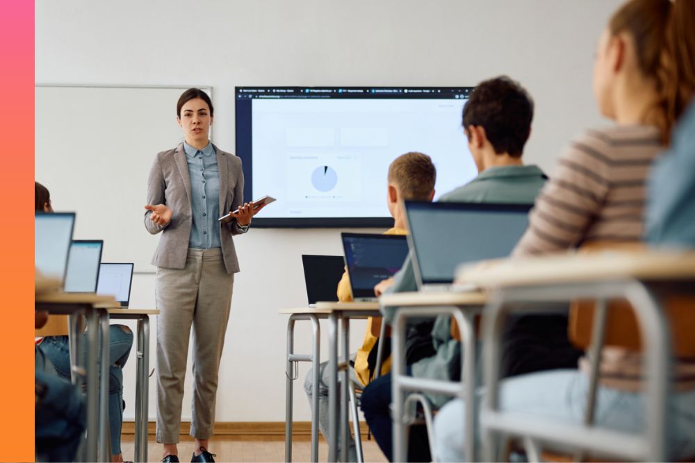 A professional stands and presents in front of a room full of people at desks with their laptops open in front of them.
