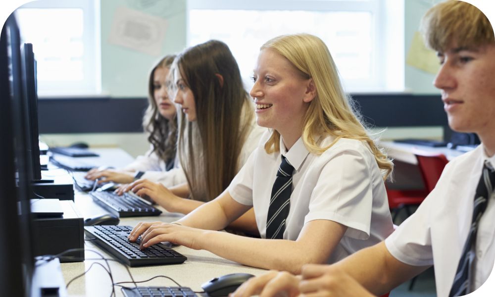 Students in uniforms sit at tables and type on keyboards connected to monitors.