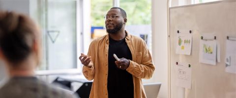 An individual presents and gestures in front of a whiteboard.