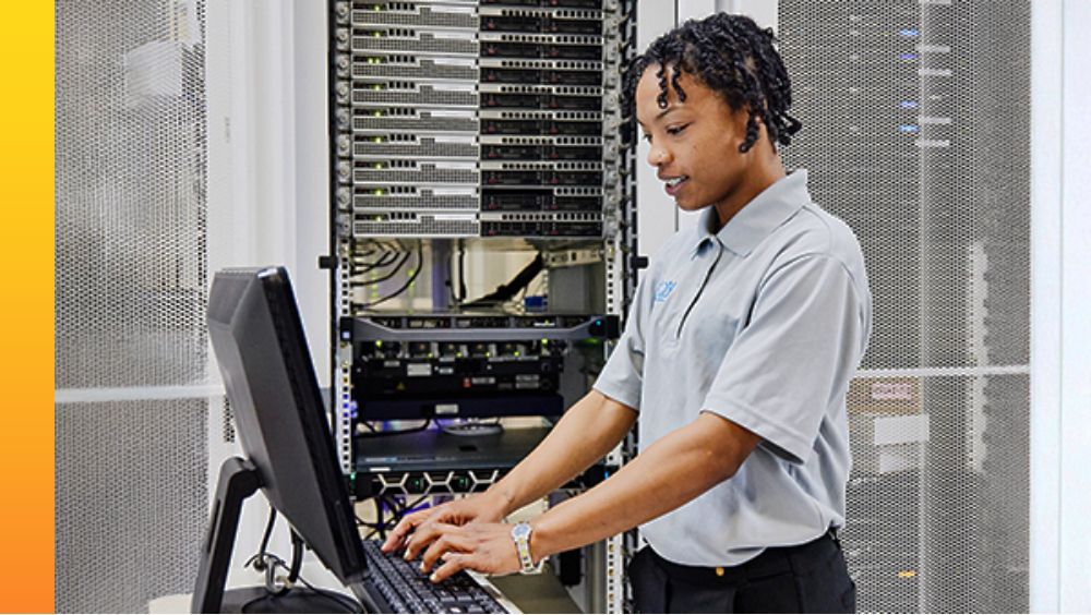 A person stands at a workstation inside a data center typing on a keyboard connected to a monitor with servers, cables and networking equipment in the background. 