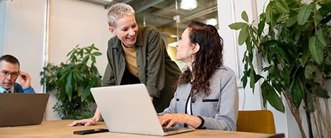 A person leans on a table to talk to another person who is seated and working on a laptop.