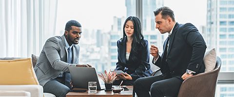 Three business professionals discuss something on a laptop in front of a window showing a cityscape 