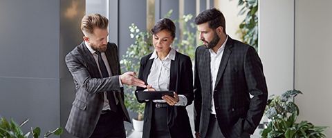Three people in business attire discuss something while referring to a tablet screen