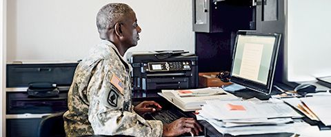 Government employee in camouflage uniform works on a desktop computer.