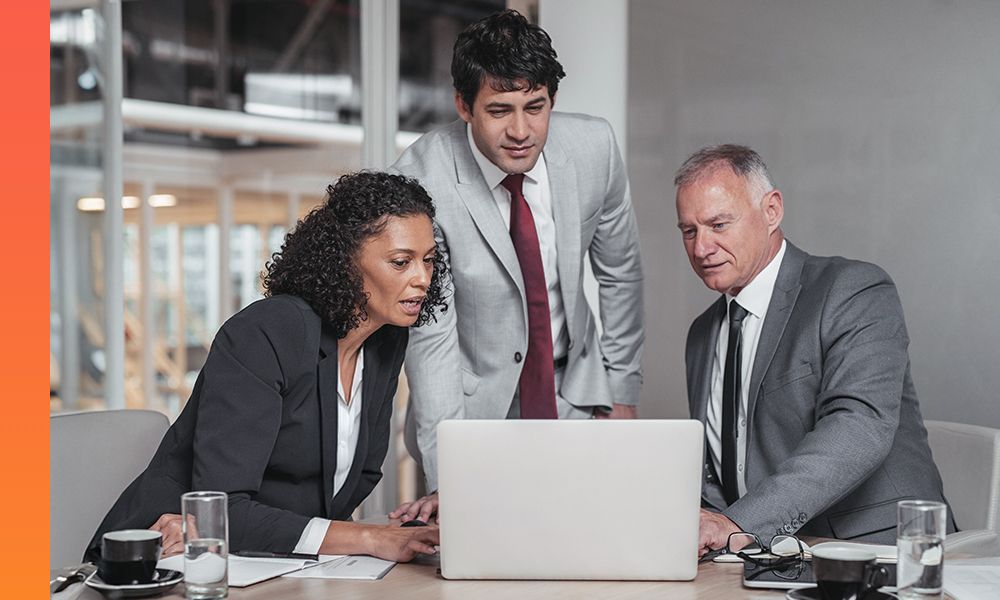 Three people in business attire discuss something while looking at a laptop
