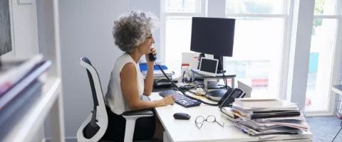 Person sitting at an office desk, speaking on a telephone while working at a computer workstation.