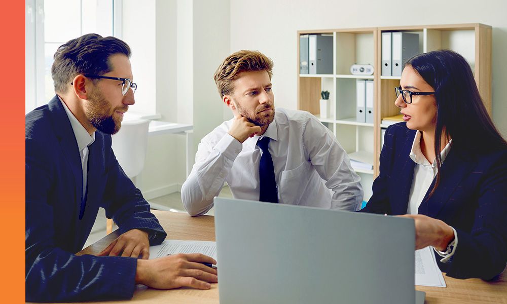 Three people seated at a conference table with an open laptop and printed documents, speaking with one another in a bright office with shelving units in the background.