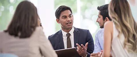 Professionals collaborating in a meeting, with one person speaking and gesturing while others listen around a conference table.