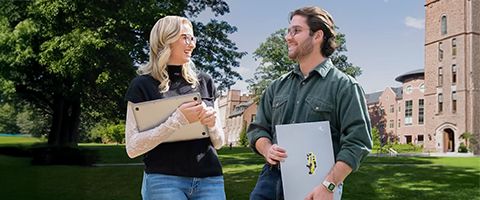 Two people carrying laptops chat together outdoors on a campus-like setting, framed by an orange graphic overlay in the background.