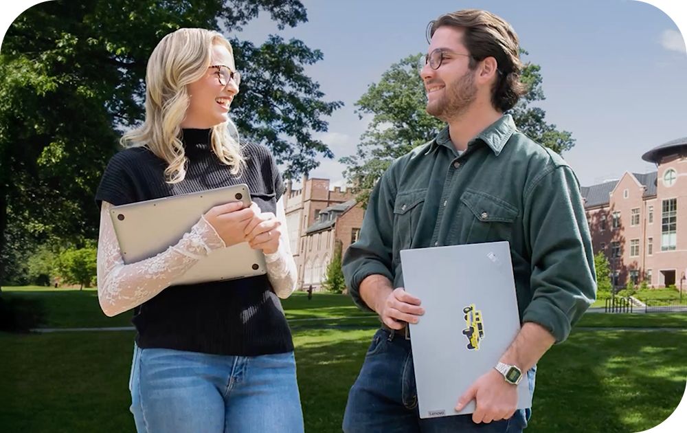 Two people carrying laptops chat together outdoors on a campus-like setting.