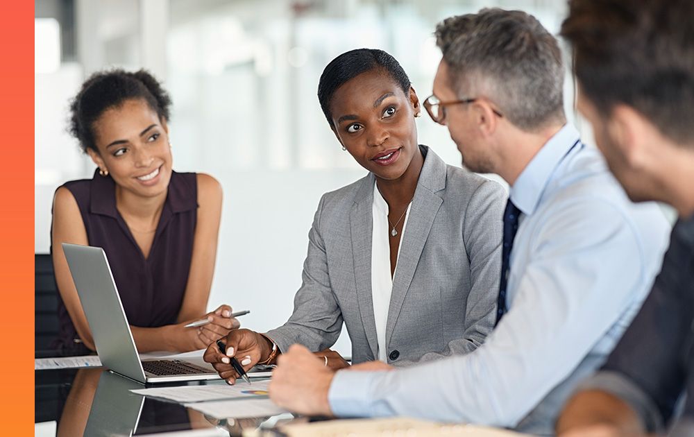 A group of four people in business attire sit and talk at a table with papers and laptops.