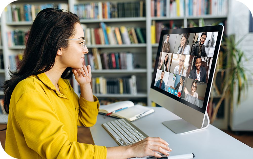 A person in a yellow top sits at a desk near a large bookshelf and participates in a video call on a desktop computer.