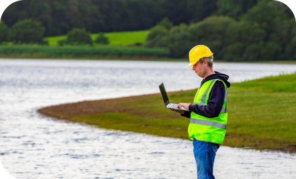 A man in a yellow safety jacket stands next to a body of water typing on a laptop.