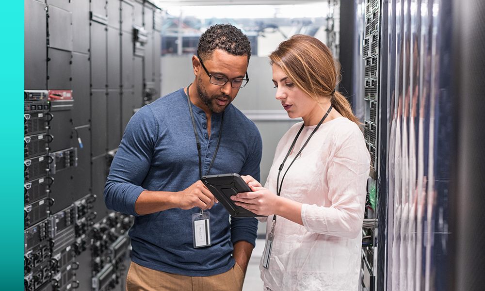 Two individuals stand between data server racks while looking at a tablet together.