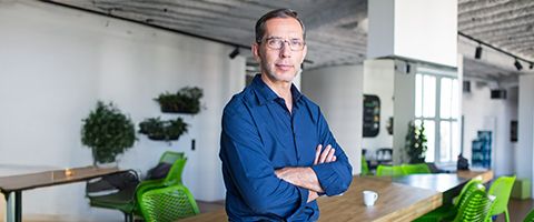 A person in a blue button-down shirt stands with their arms crossed in a bright, modern office.