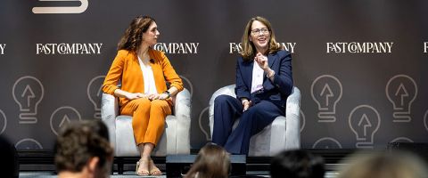Two women in business suits sitting on an elevated stage while giving a talk in front of a crowd of people 