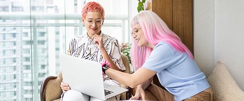 Two businesspeople are in conversation while gesturing at a laptop in one of their laps.
