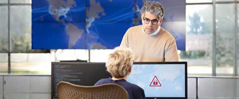 Two businesspeople are in conversation, with one leaning over the other’s monitor. There is a world map displayed over multiple screens in the background. 
