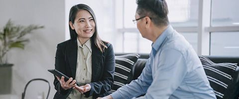 Two businesspeople sat on the couch in conversation with one holding a tablet. 