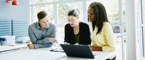 Three professionals huddle together at a table in front of a tablet to communicate seamlessly