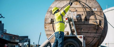 A Lumen worker in yellow hardhat and safety vest unhooks a wooden fiber roll from a utility truck