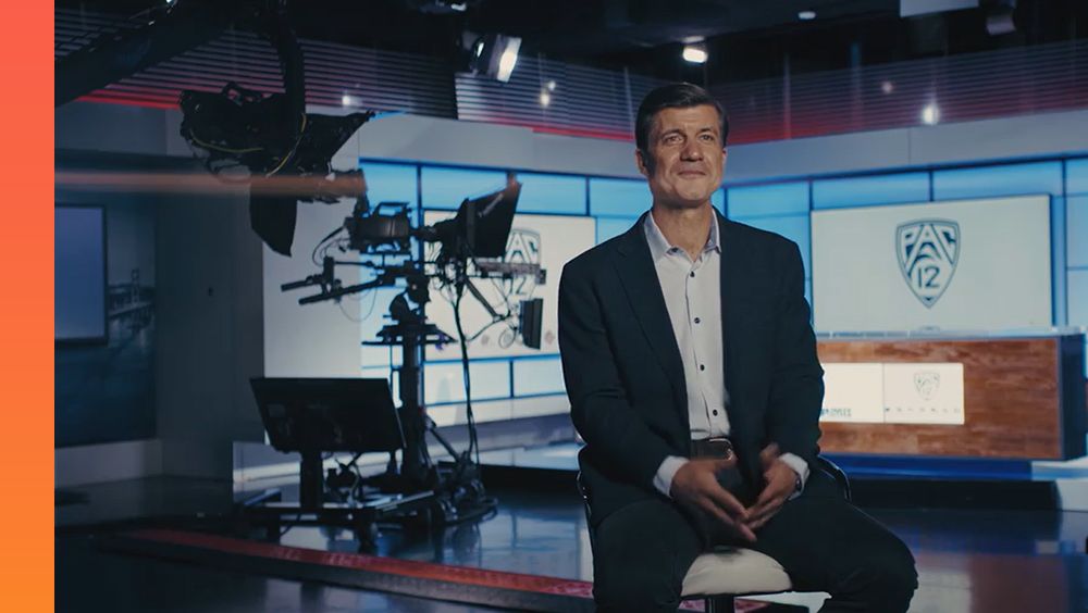 Person in a suit talks into a camera while being interviewed in a studio with Pac 12 logo on screens in the background.