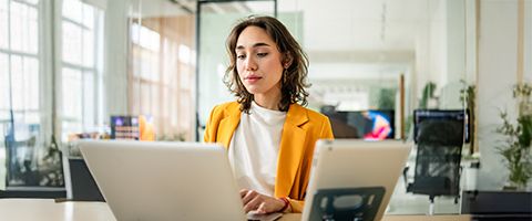 A person in a yellow blazer works at a laptop on a desk with a tablet propped up close by.