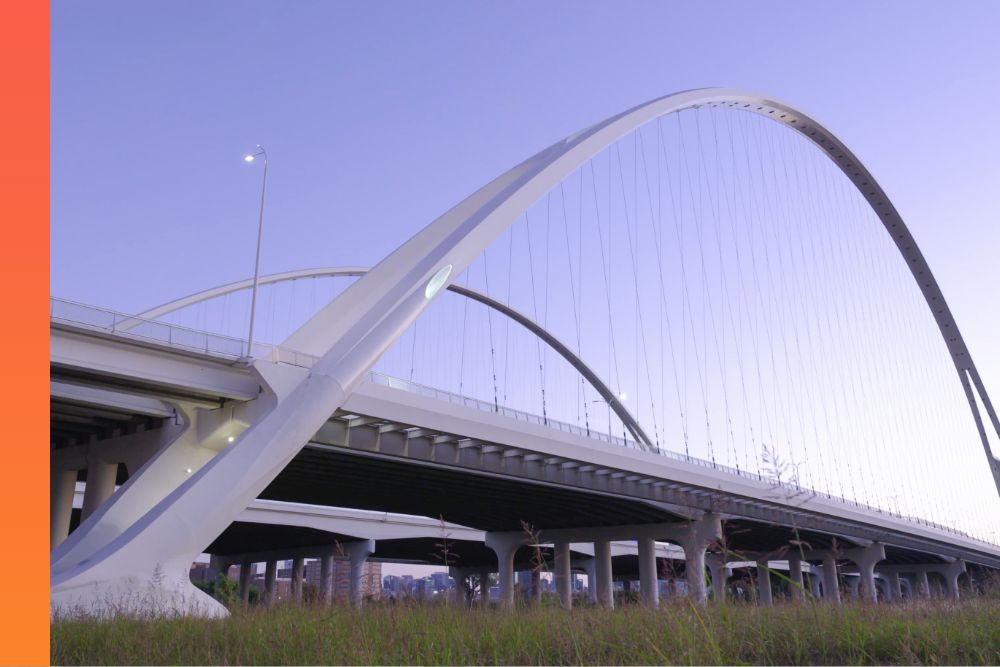 Modern arched bridge with sweeping white steel supports and suspension cables, viewed from below against a clear, pale blue sky. 