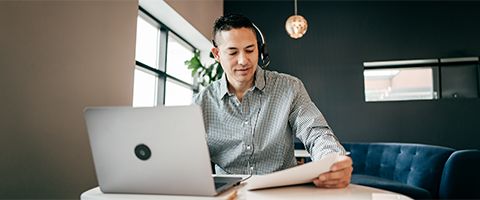A professional wearing a headset and working on a laptop examines some papers.