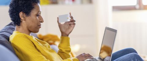 A person sitting on a couch holding a credit card in one hand and using a laptop to browse the internet with the other hand.