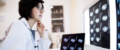 A medical professional wearing a white views images of a human brain on light board and computer screen in an office with medical supplies on shelves in the background. 