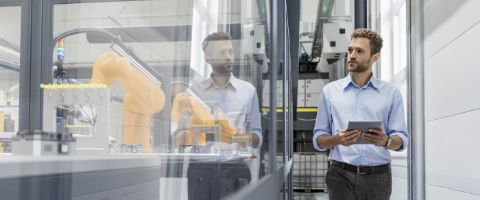A person in a high-tech manufacturing environment holding a tablet views a robotic arm and other pieces of industrial equipment and machinery through a glass window.
